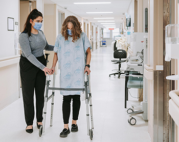 Aide soignante qui accompagne une femme marchant à l'aide d'une marchette.