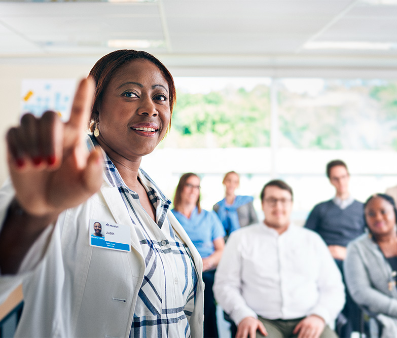 Professionnelle de la santé qui pointe le doigt sur la caméra avec en arrière plan un groupe assis